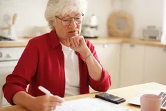 Seniorka v červeném svetru počítá domácí rozpočet u stolu s papíry a kalkulačkou