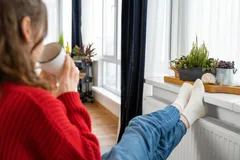 Žena v červeném svetru relaxuje doma s šálkem čaje u okna s květinami