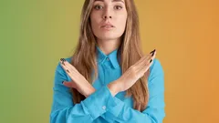 Žena v modré košili gestikuluje znak X rukama na barevném pozadí, vyjadřuje nesouhlas.