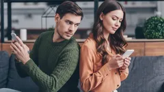 Žárlivý muž sleduje ženu píšící na mobilním telefonu, oba sedí zády k sobě na gauči.