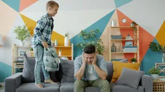 Otec ve stresu s bolestí hlavy, syn skáče na pohovce a bije ho polštářem